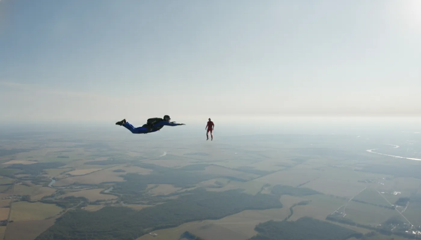 A student skydiver in a flat tracking position moving across the sky, arms swept back, a single instructor visible some distance away against a bright hazy horizon