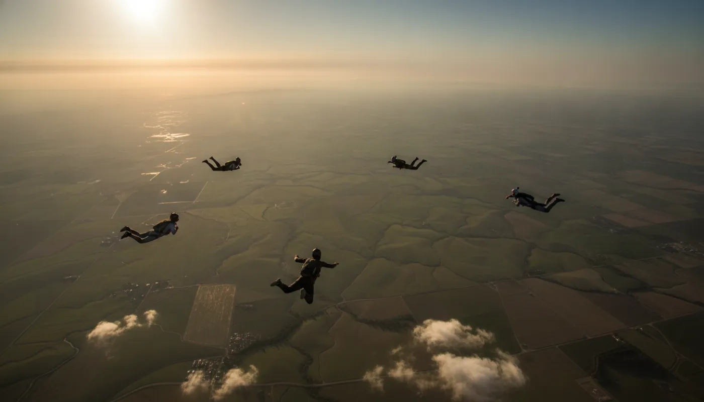 A four-way belly formation breaking off over patchwork farmland far below, jumpers tracking away from each other in the late-day haze