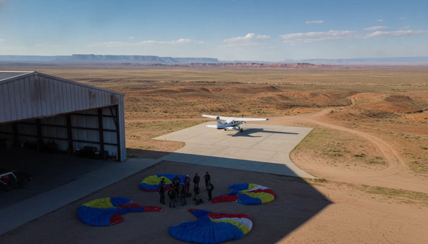 Skydivers gathered in the shade beside an open hangar door, rigs laid out on packing mats while a small aircraft waits on the apron beyond, midday desert light