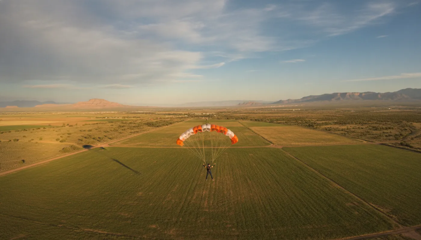A skydiver under a large docile main canopy on final approach to a grass landing area, arms up before the flare, long shadows across the field