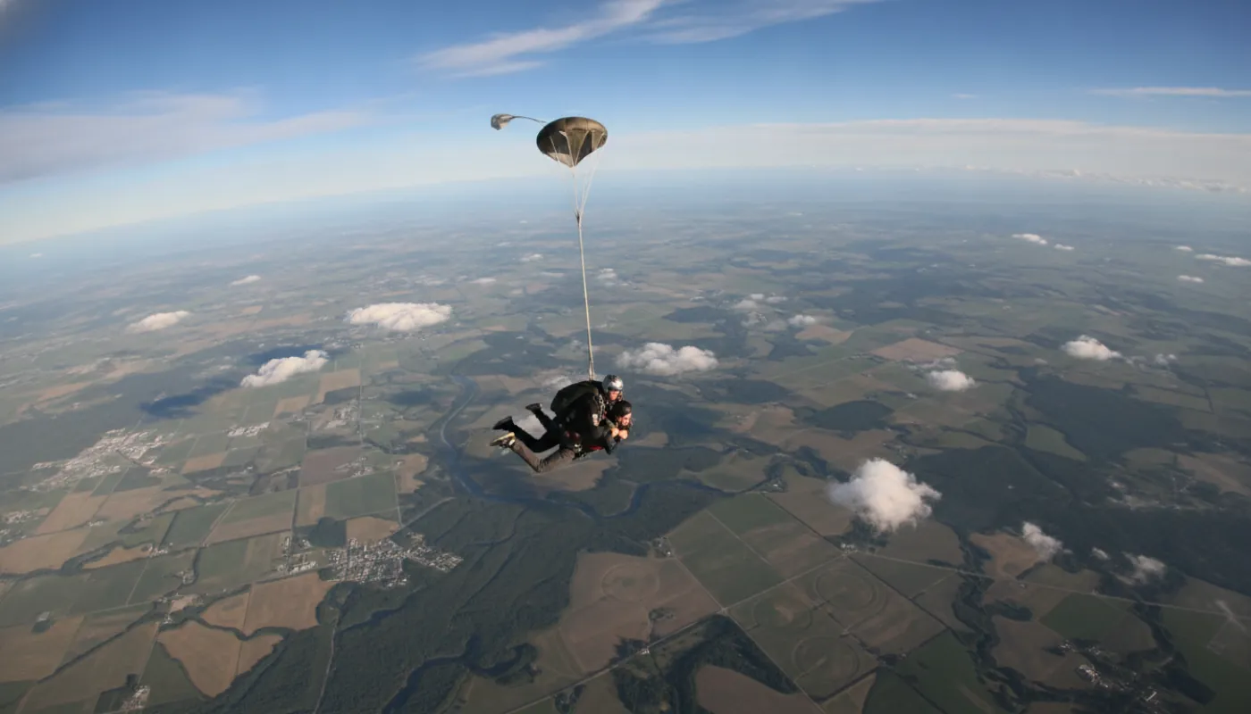 A tandem pair moments after exit, arms crossed and heads back, the drogue chute streaming above them against a wide patchwork of fields far below