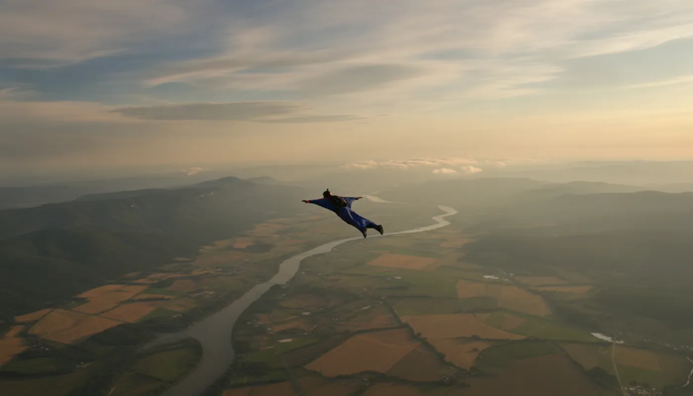 A lone wingsuit pilot in flight high above a broad river valley, arms and legs spread taut, patchwork fields and a distant ridgeline far below in hazy afternoon light